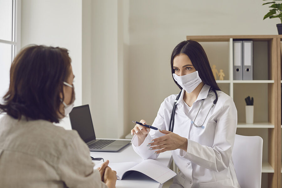 Female doctor and patient in protective masks talking at clinic. Client receiving consultation from medic at hospital