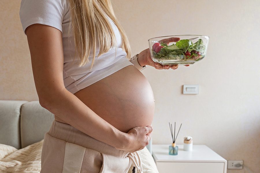 Happy young pregnant woman eating vegetable salad at home. Healthy nutrition and pregnancy concept. Expecting mother holding bowl with fresh cucumbers and tomatoes