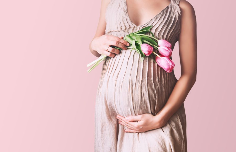 Beautiful pregnant woman with tulips flowers holds hands on belly in white background. Young woman in maternity dress waiting for baby birth. Pregnancy, Motherhood, Mother's Day Holiday concept.